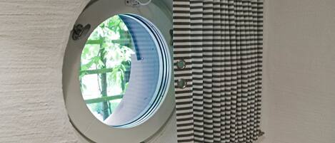 Una ventana circular con vistas a la vegetación, una cortina a rayas y una pared blanca con diseño de ladrillo.