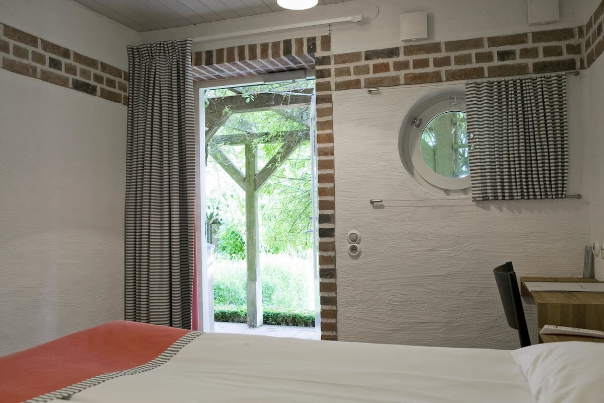 Un dormitorio con cama, una ventana con cortinas, un espejo redondo y una mesita de noche de madera.