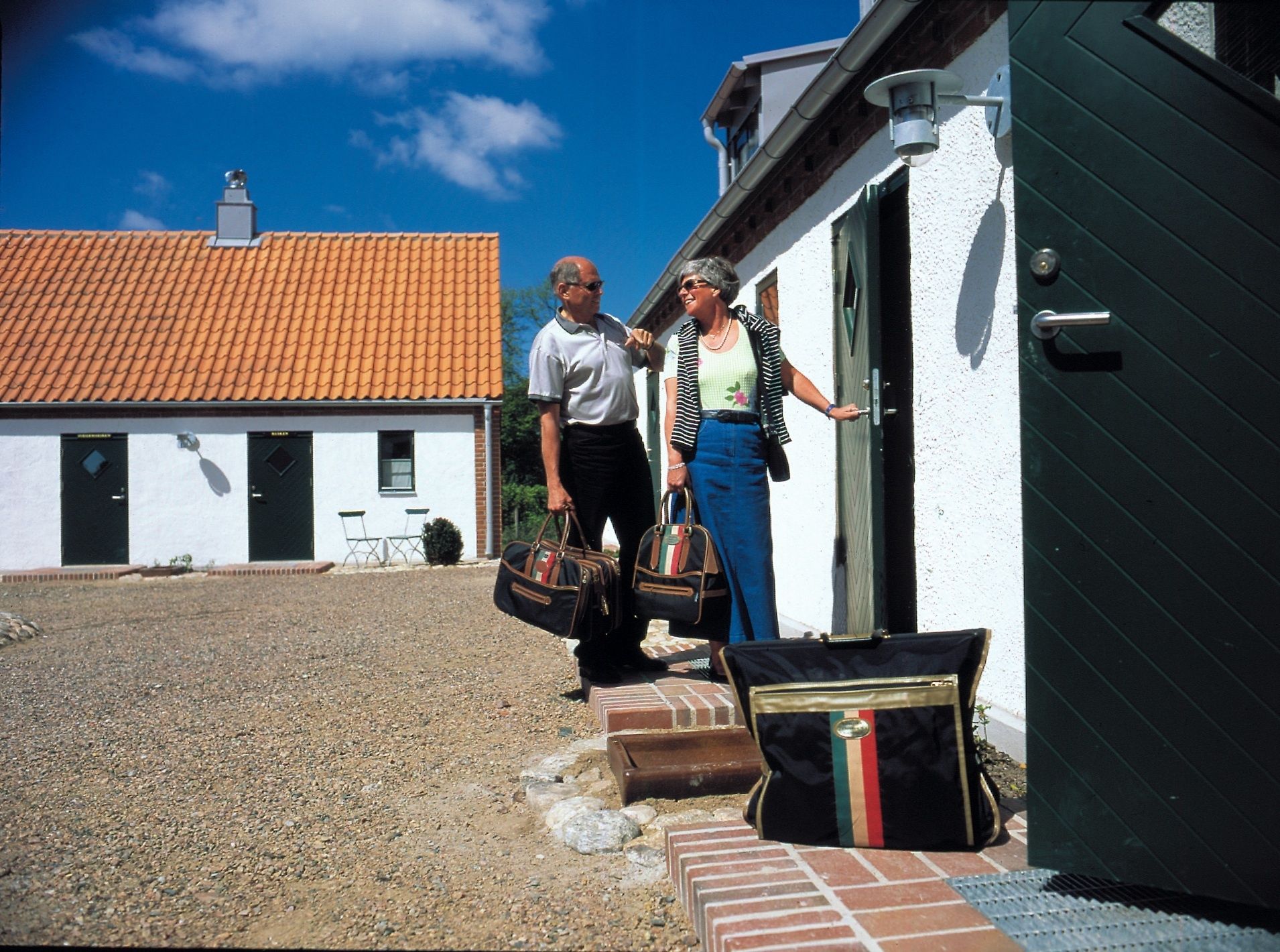 A building with a white exterior and a red-tiled roof. There are two people standing at the entrance, one holding a bag.