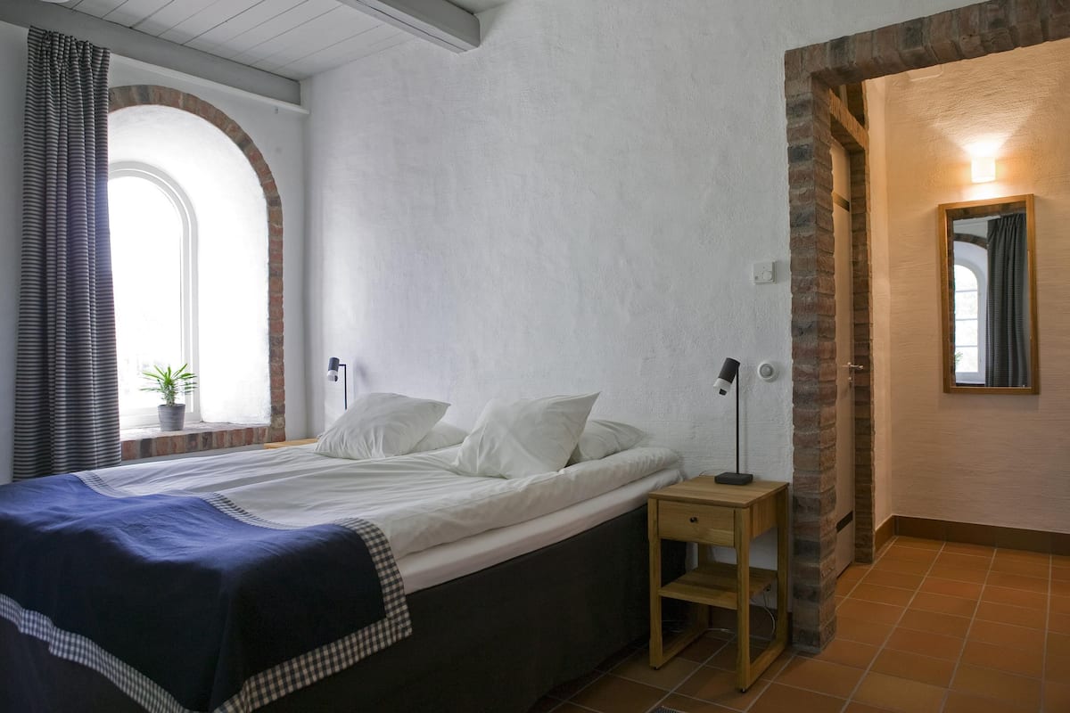 Un dormitorio con cama, mesita de noche, lámpara, ventana con cortinas y una planta en maceta.