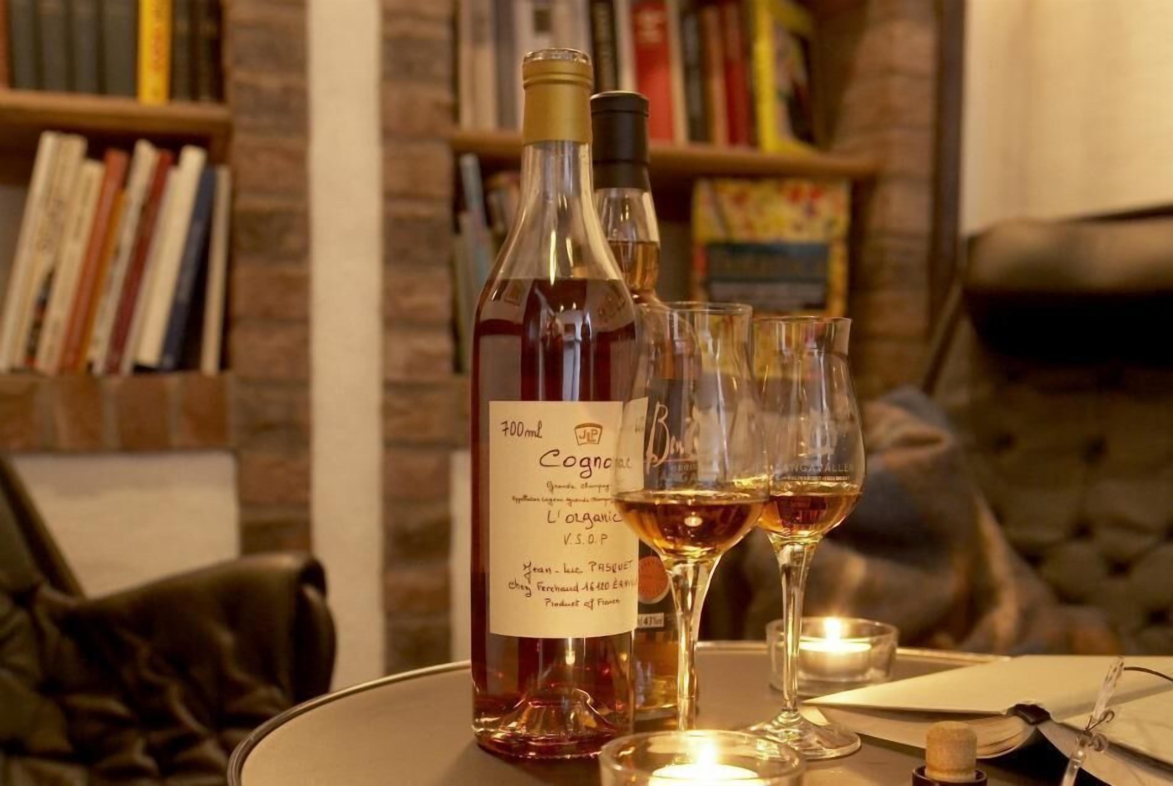 En flaske cognac og to glas på et bord med et lys, bogreoler og en lædersofa i baggrunden.