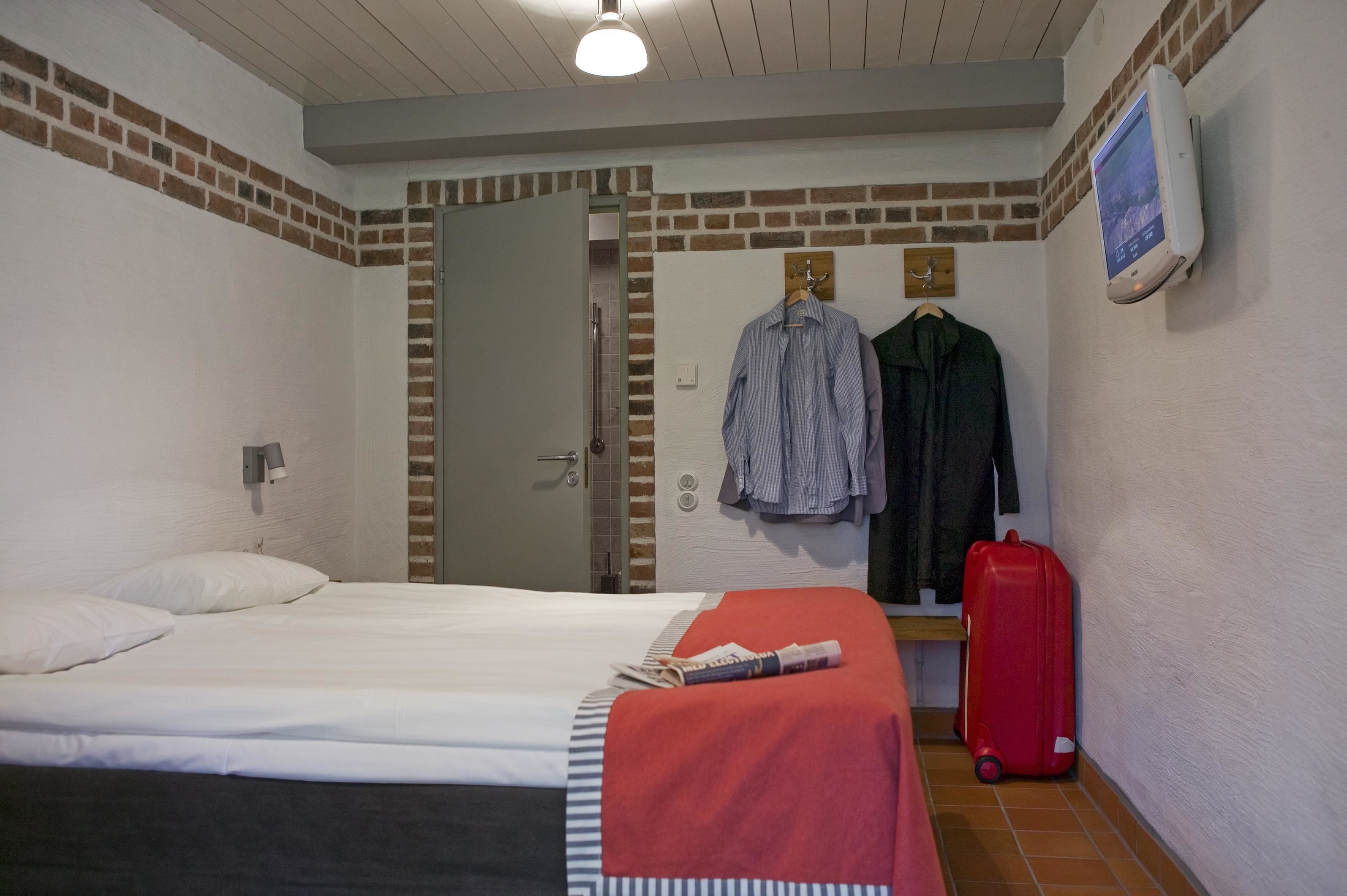 Ett hotellrum med en säng, en röd resväska och en klädställning med två jackor.