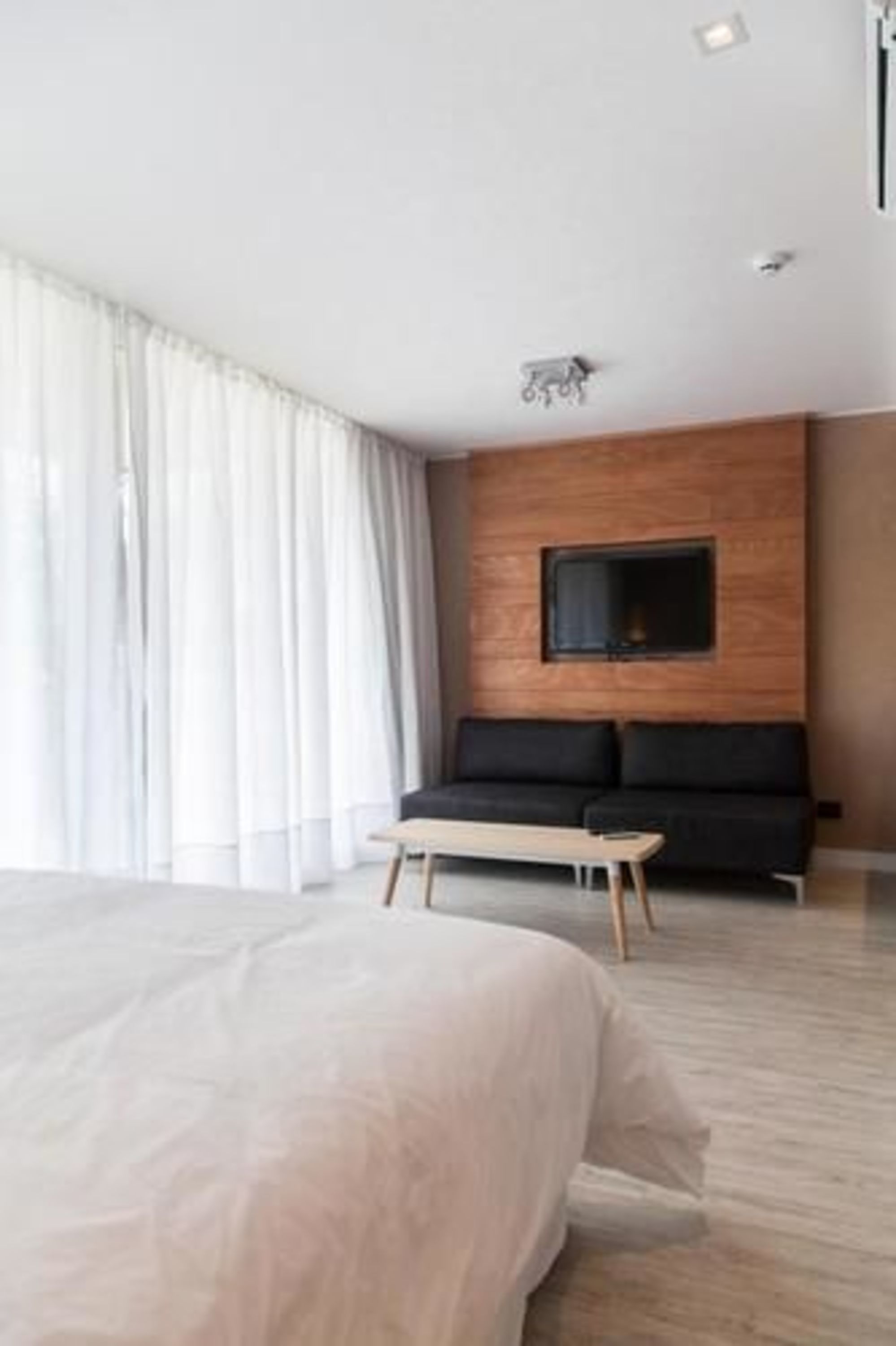 A modern hotel room with a large bed, a wooden accent wall, a flat-screen TV mounted above a black sofa, and a large window with sheer curtains.