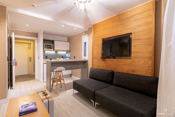 A modern living room with a grey sofa, a wooden accent wall, a mounted TV, and a kitchen area with a microwave. - 2122 Hotel Art Design (Punta del Este)