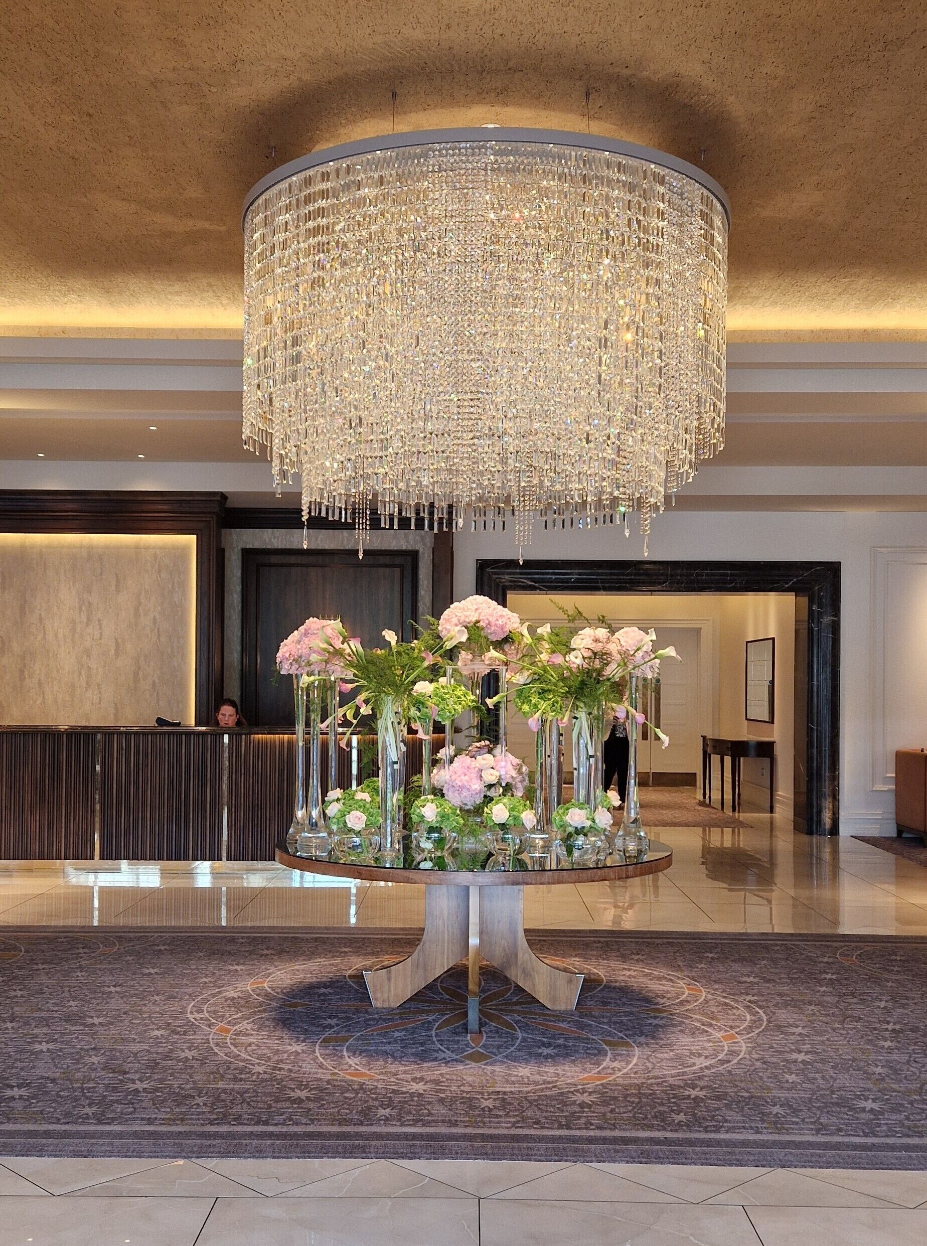 A lobby with a large chandelier, a reception desk, and a round table with floral arrangements.