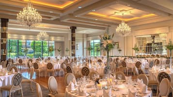 Un salón de banquetes con varias mesas redondas, candelabros y ventanas amplias que dan a zonas verdes.