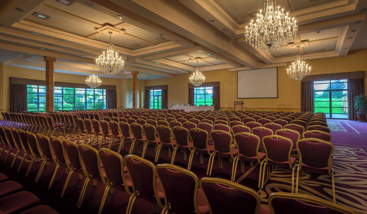 Un salón de conferencias con filas de sillas, candelabros, una pantalla de proyector y ventanas amplias.
