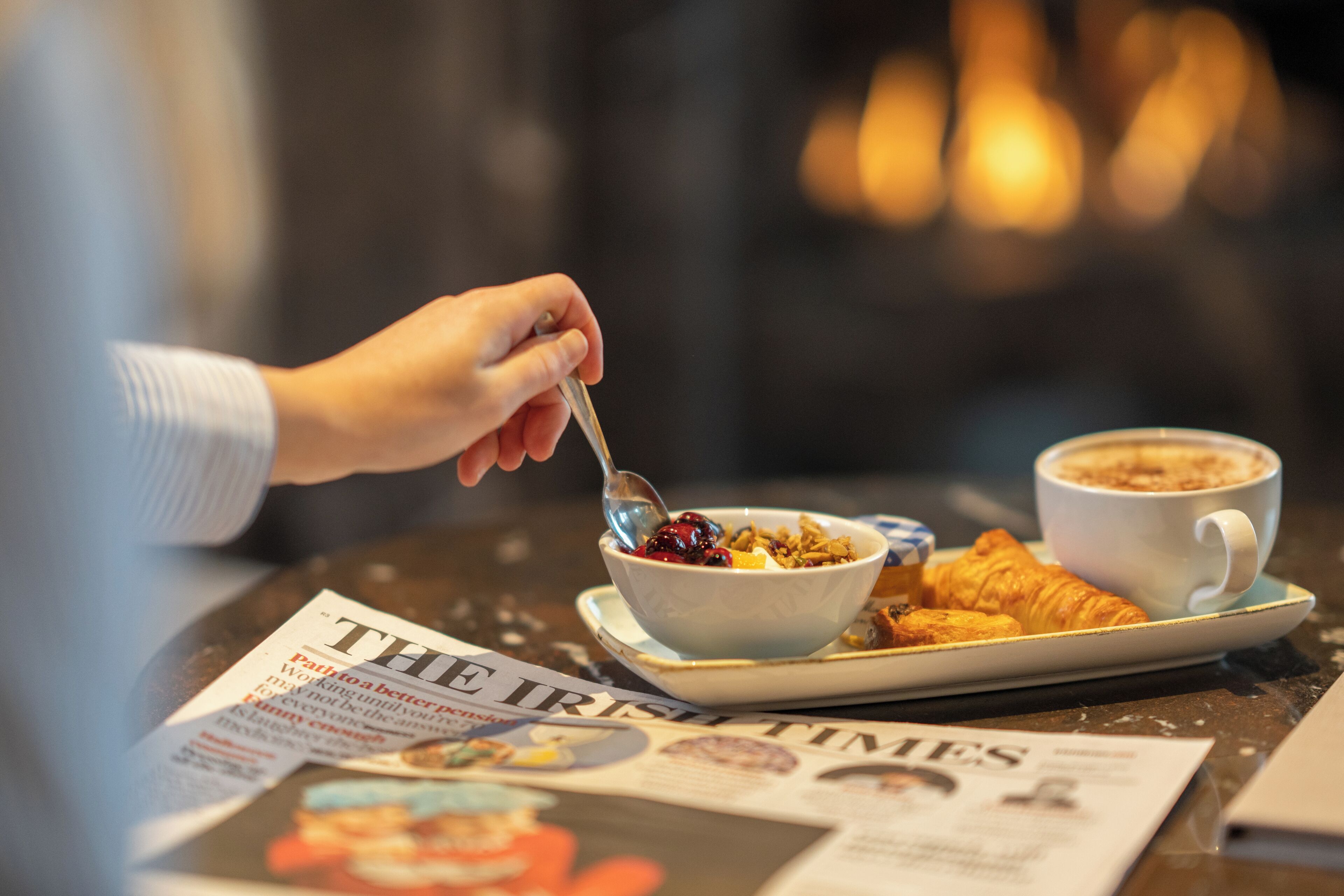 A breakfast setting with a newspaper, a bowl of cereal with fruit, a croissant, and a cup of coffee on a table.