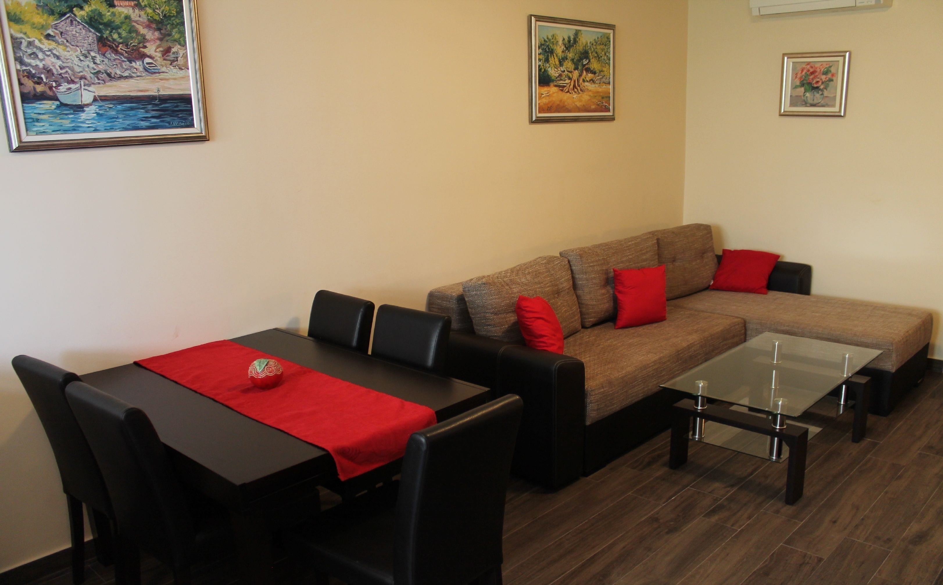 A living room with a brown sofa, black chairs, a glass coffee table, and a red table runner. There are three framed artworks on the wall.