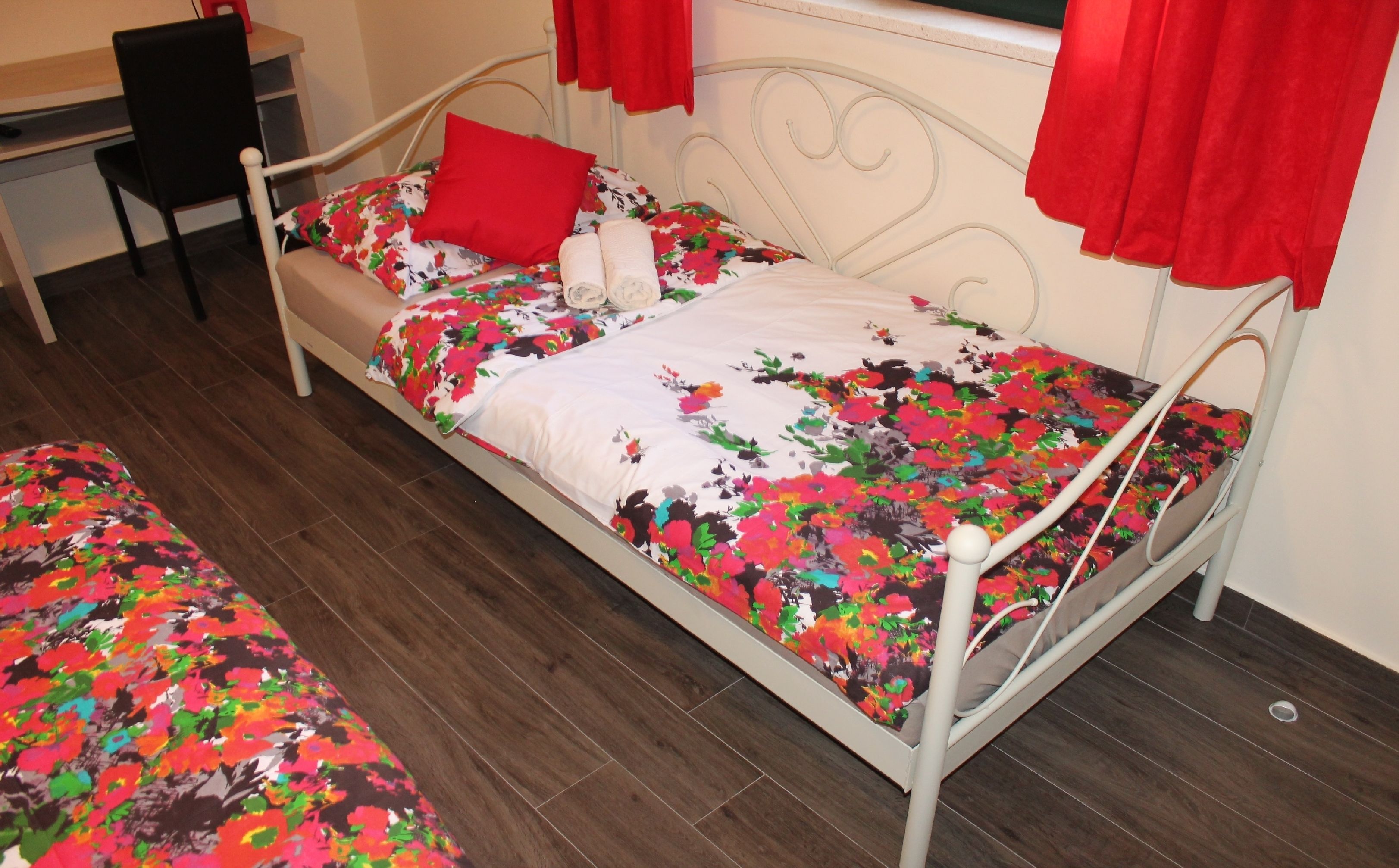 Ein Einzelbett mit einem blumigen Überwurf, einem roten Kissen und einem weißen Kissen. Neben dem Bett steht ein Stuhl und im Hintergrund ein Schreibtisch.