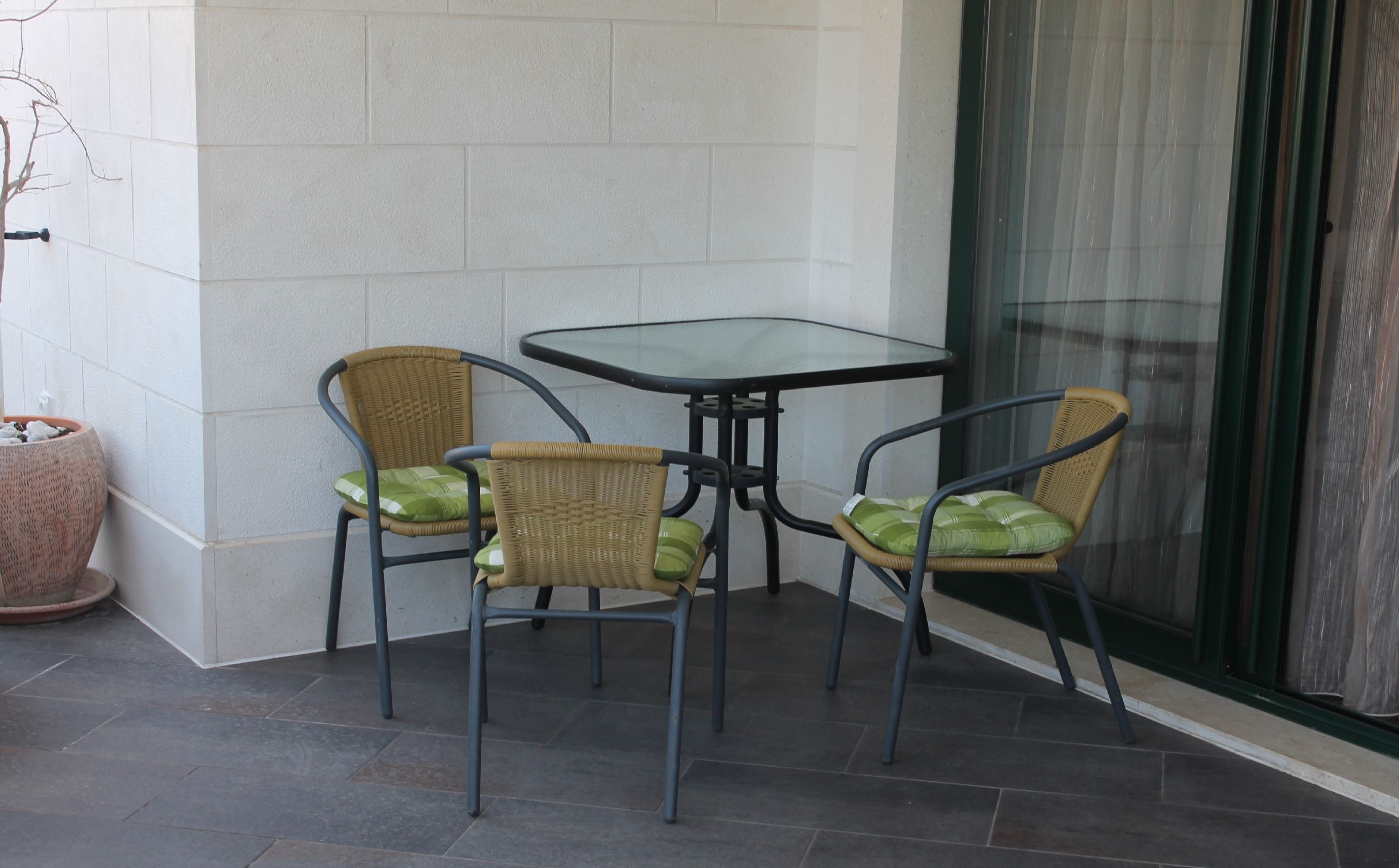 Een hoekterras met een glazen tafel en twee rieten stoelen met groene kussens.