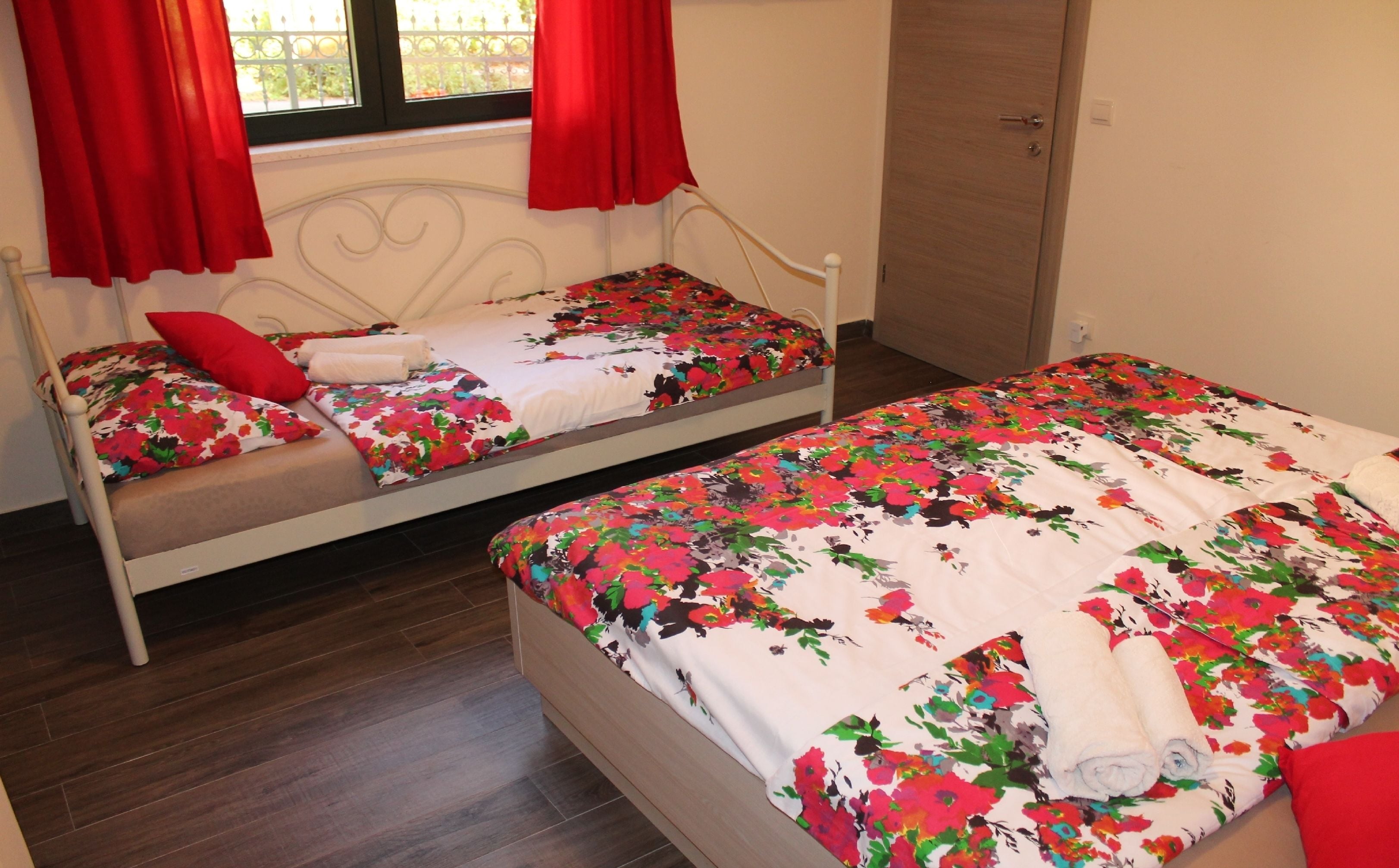 Dos camas individuales con ropa de cama floral, un armario de madera y una ventana con cortinas rojas.