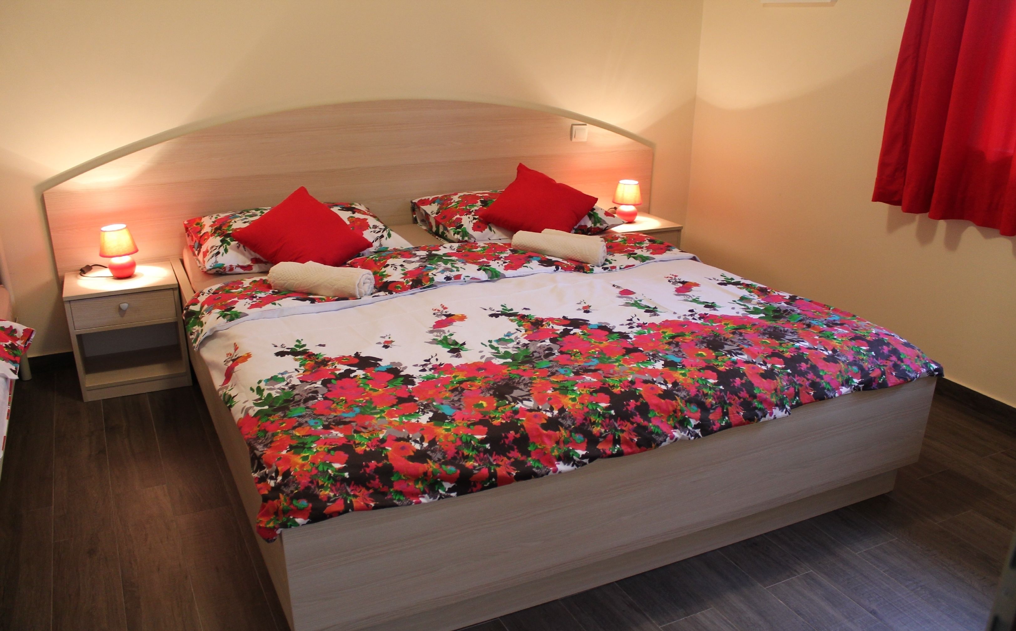 Una cama con ropa de cama floral, dos almohadas rojas y una lámpara de noche a cada lado.