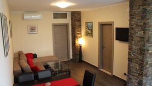 A modern living room with a sofa, chairs, a glass coffee table, and a TV mounted on the wall. There are two doors, one with a stone accent wall, and an air conditioning unit above the sofa. - Apartments Logoš Cavtat (Konavle)