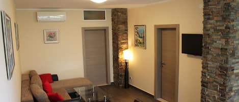 A modern living room with a sofa, chairs, a glass coffee table, and a TV mounted on the wall. There are two doors, one with a stone accent wall, and an air conditioning unit above the sofa.