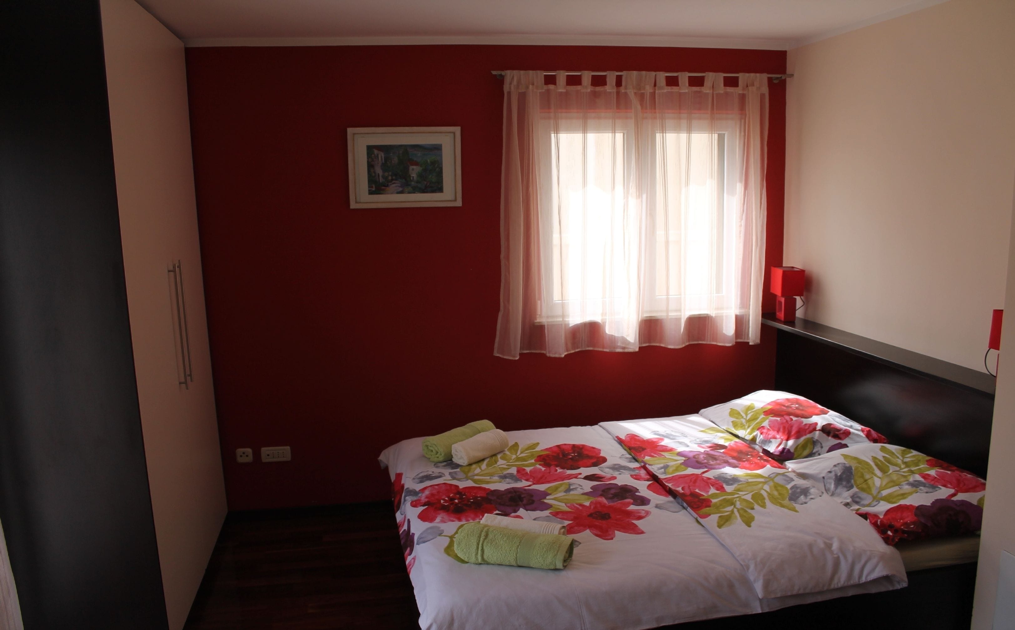 A bedroom with a bed, a window with sheer curtains, a painting on the wall, and a red bedside table.
