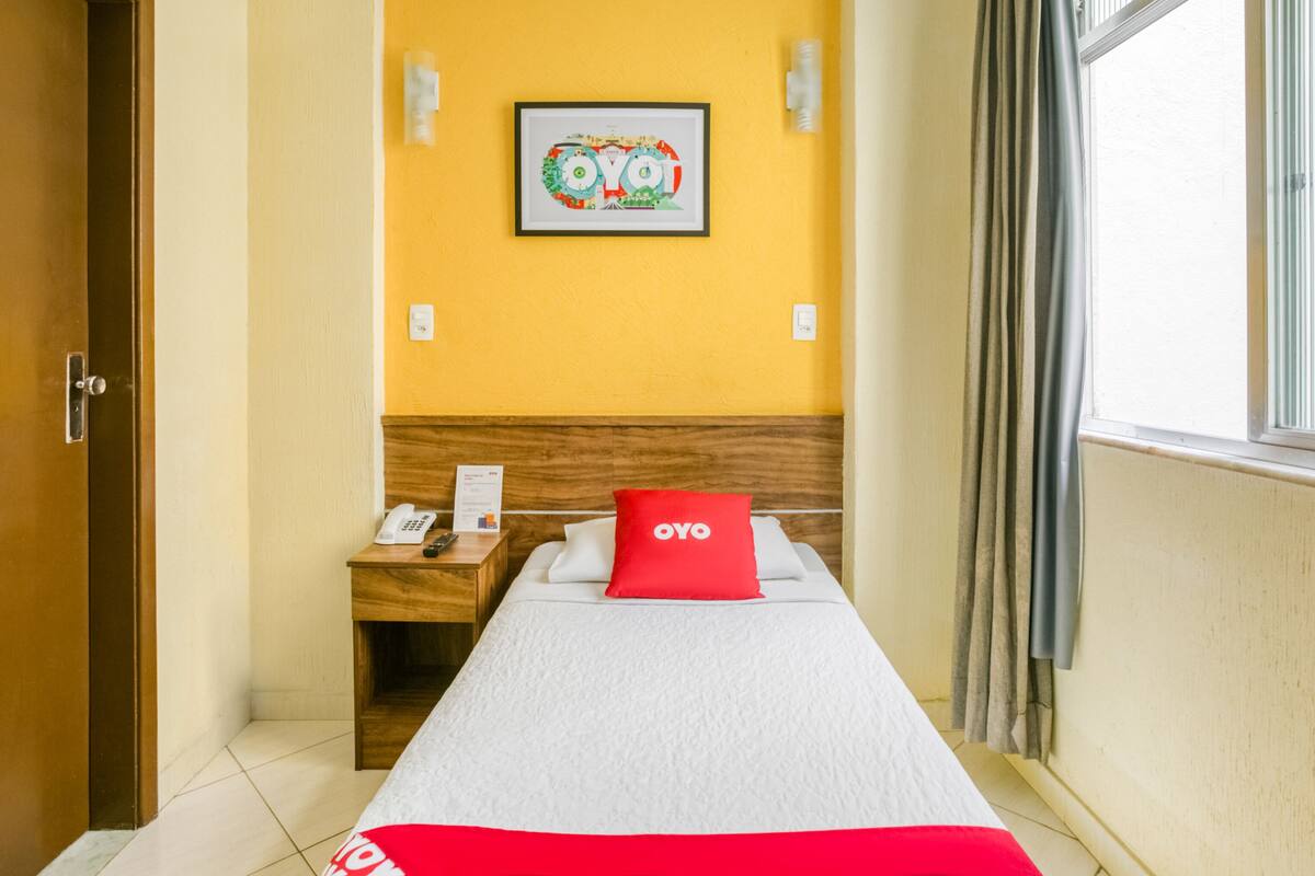 Una habitación de hotel con cama, mesita de noche, teléfono, un cuadro en la pared y una ventana con cortinas.