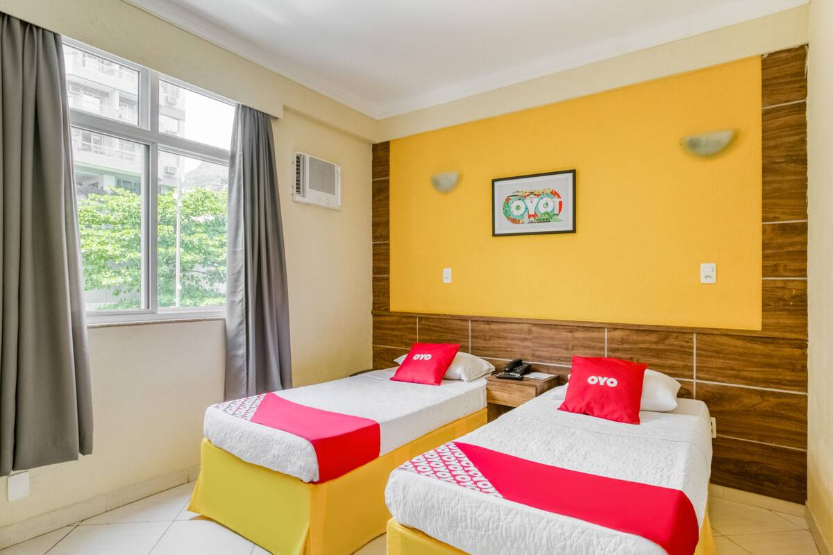 Una habitación de hotel con dos camas, una ventana con cortinas, un aire acondicionado instalado en la pared y un cuadro enmarcado.