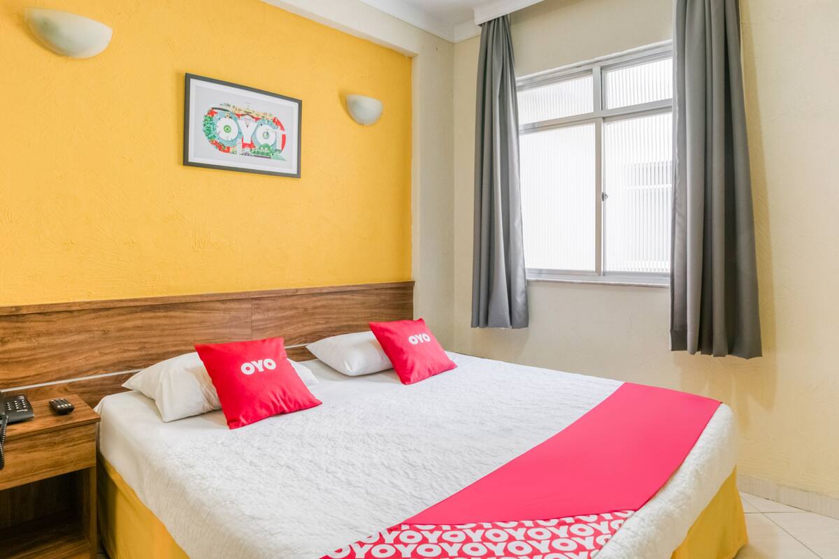Una habitación de hotel con cama, mesita de noche, un cuadro en la pared y una ventana con cortinas.