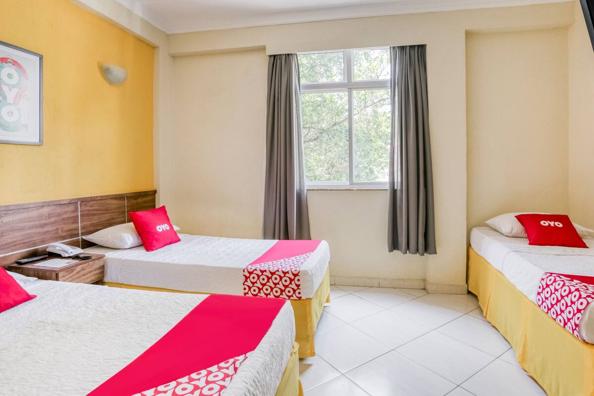 Habitación de hotel con dos camas, cada una con una colcha de cuadros rojos y blancos, un cabecero de madera, una mesita de noche con teléfono y control remoto, y una ventana con cortinas grises.