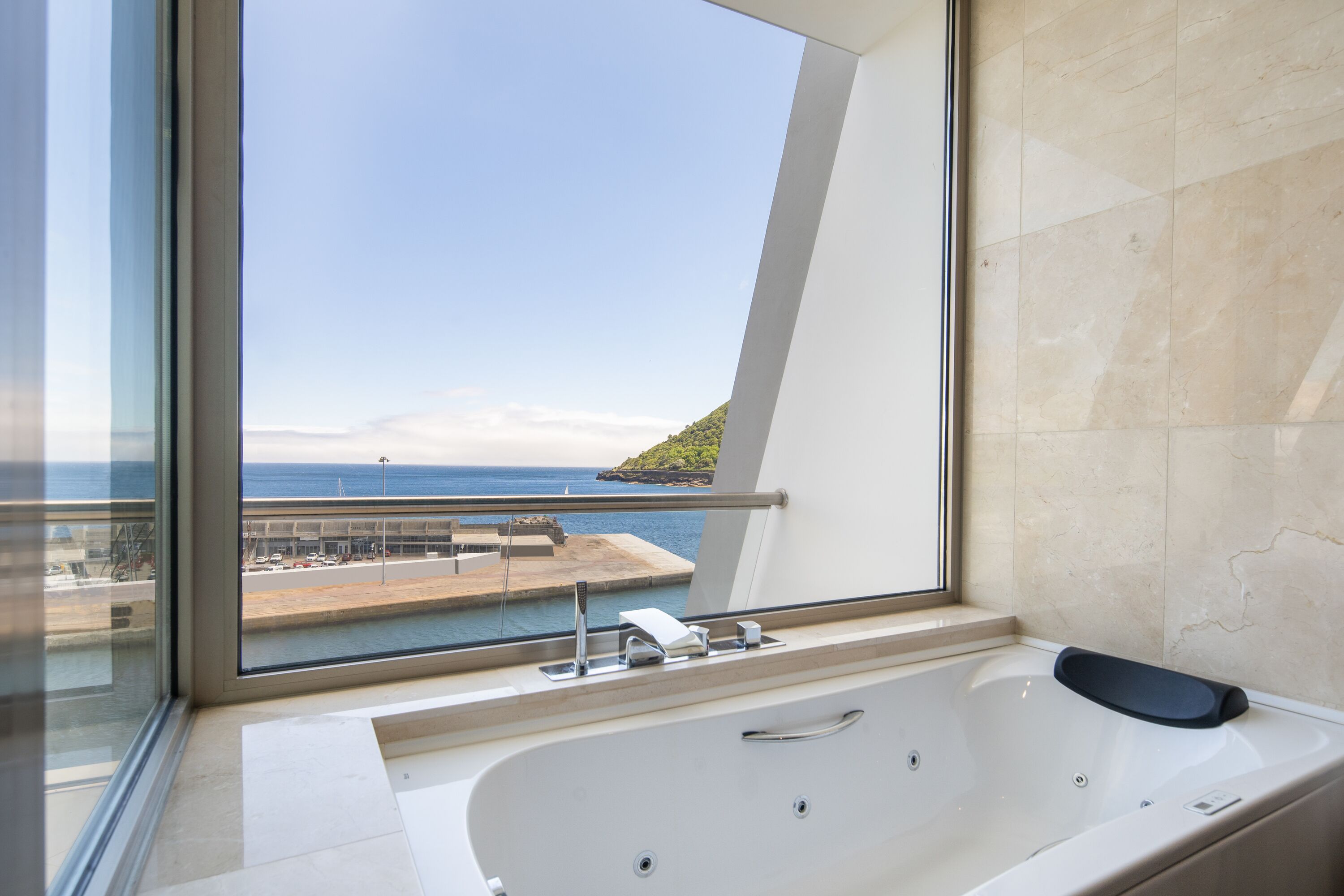 A modern bathroom with a large bathtub, a window with a view of the sea and a distant island, and a marble-tiled wall.