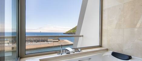 A modern bathroom with a large bathtub, a window with a view of the sea and a distant island, and a marble-tiled wall.