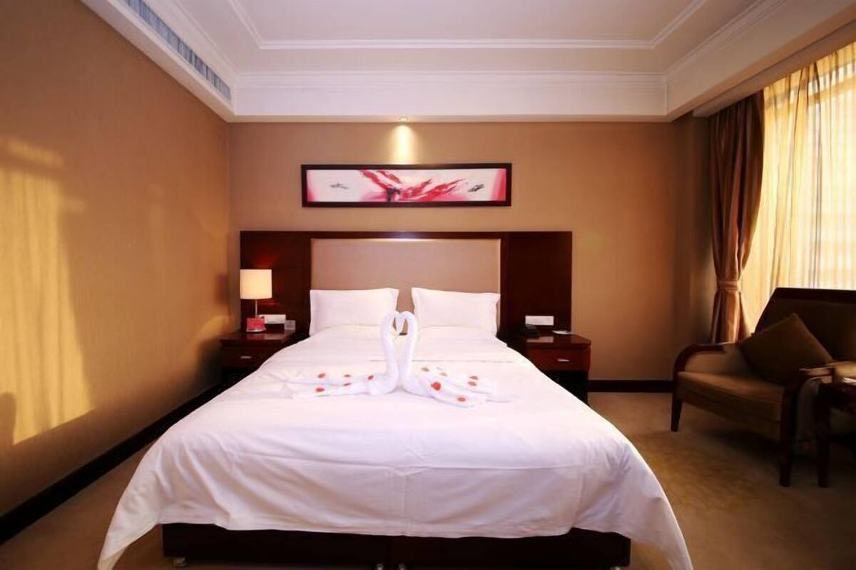 Une chambre d’hôtel avec un grand lit, une tête de lit ornée d’une œuvre d’art encadrée, une table de chevet avec une lampe et une chaise.