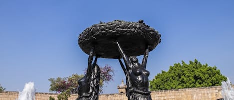 Una fuente con una escultura central y chorros de agua, rodeada de arcos de piedra y follaje.