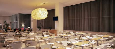 A spacious dining area with multiple tables and chairs, a large chandelier, and a television screen on the wall.