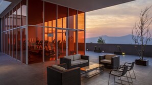 Un área de estar moderna al aire libre con mobiliario de mimbre y una partición de vidrio, con vistas a un paisaje montañoso al atardecer.