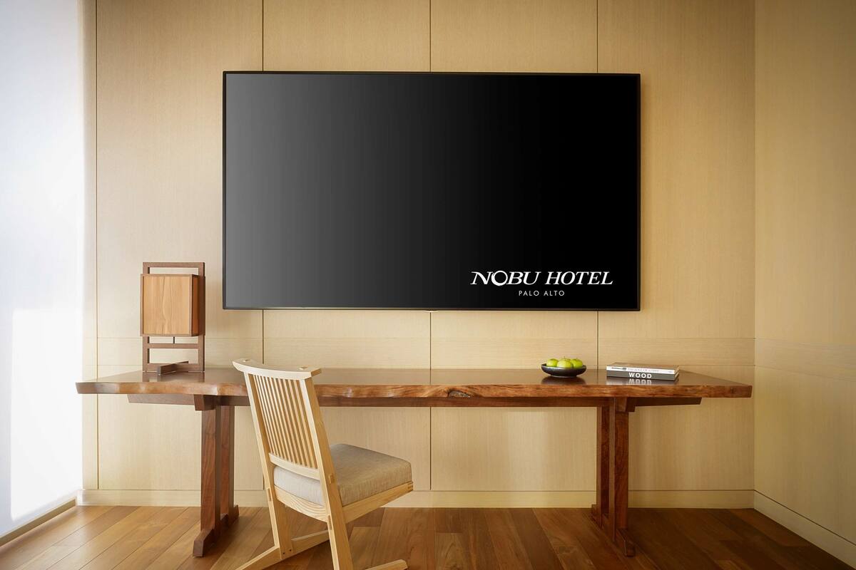 A modern hotel room with a wooden desk, a flat-screen TV mounted on the wall, a wooden rocking chair, and a small table with a bowl of fruit and a book.