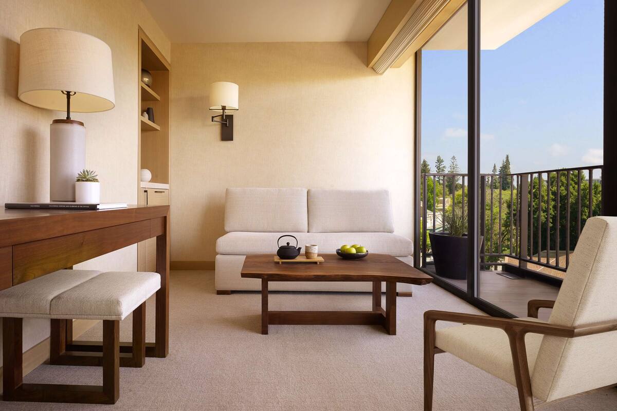 A modern living room with a sofa, a wooden coffee table, a bench, a desk with a lamp, and a view of greenery through large windows.