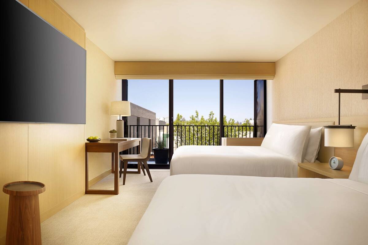 Habitación de hotel con dos camas, un escritorio, una silla, una mesita y una planta en maceta, con vistas al exterior.