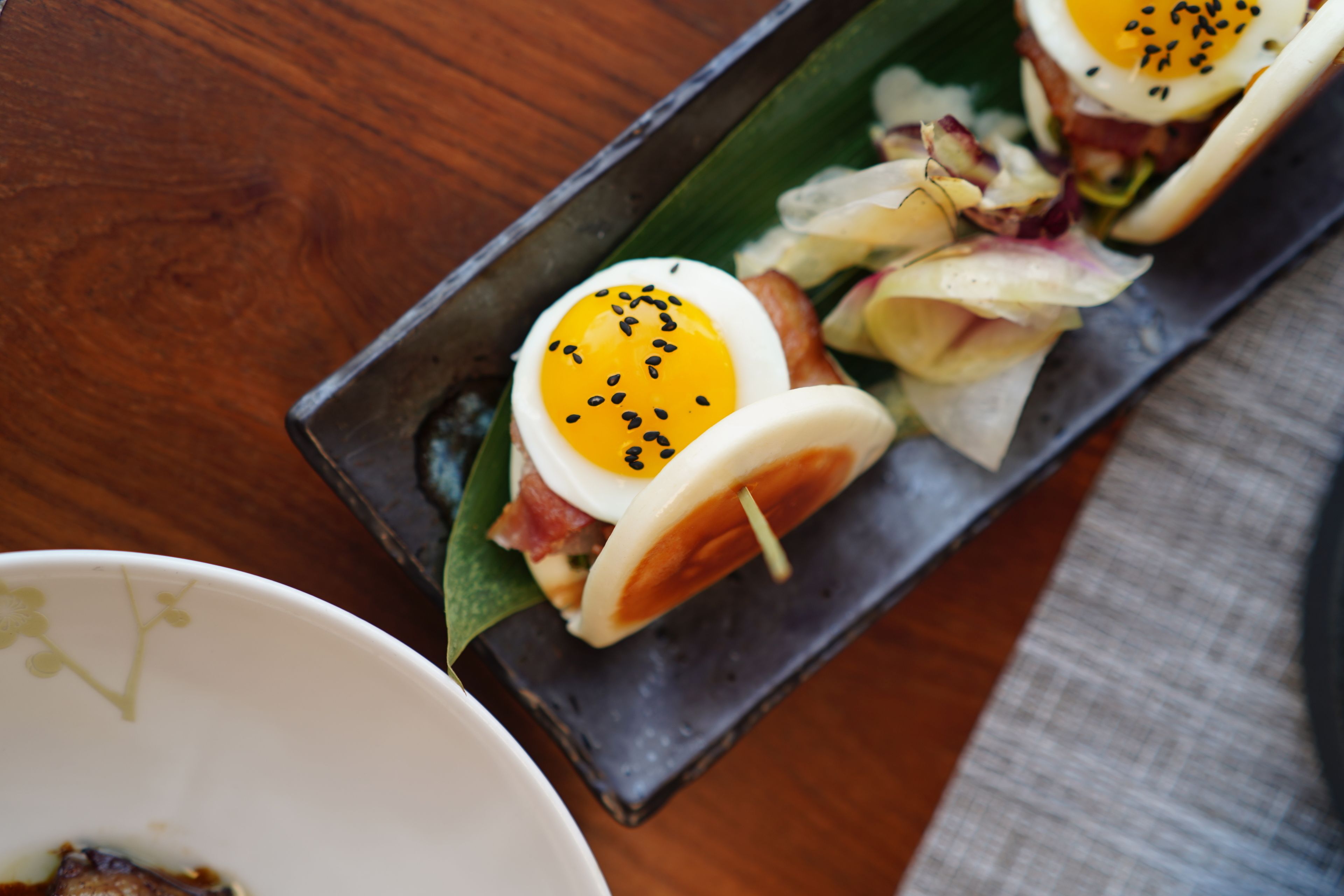 A close-up of a dish with a poached egg on top, garnished with black sesame seeds, served on a rectangular plate.