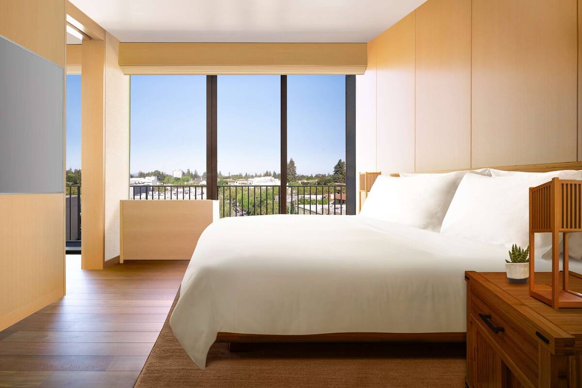 A modern bedroom with a large bed, wooden nightstands, and a view of the city through floor-to-ceiling windows.