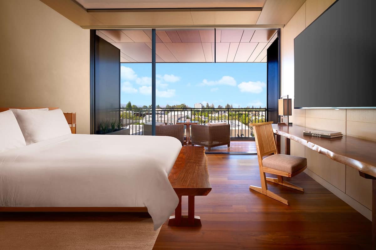 Habitación de hotel moderna con una cama grande, un escritorio de madera, una silla y un balcón con vistas a la ciudad.