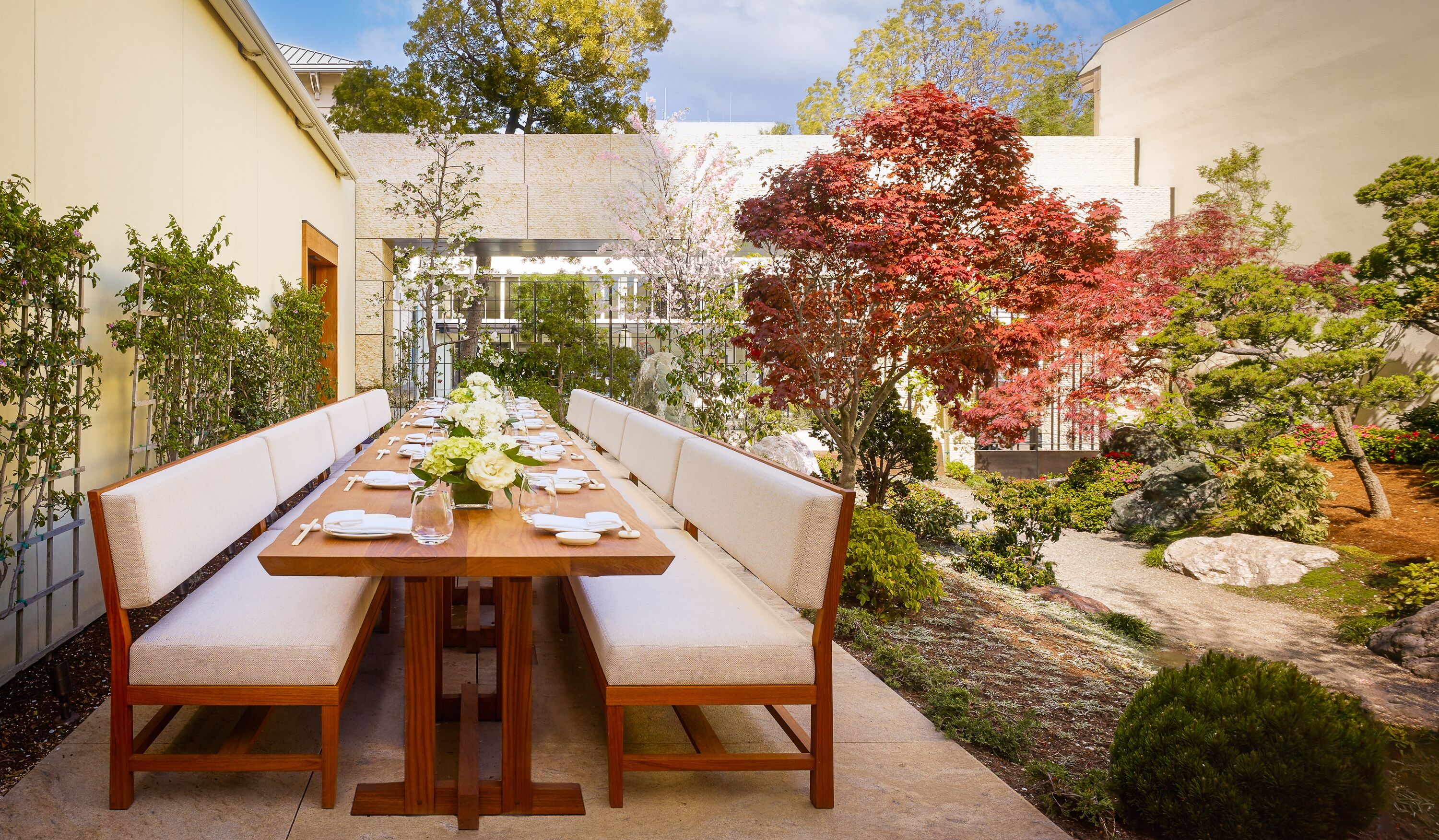 A long wooden table set for dining with white cushioned benches, surrounded by a garden with various plants and a red-leaved tree.