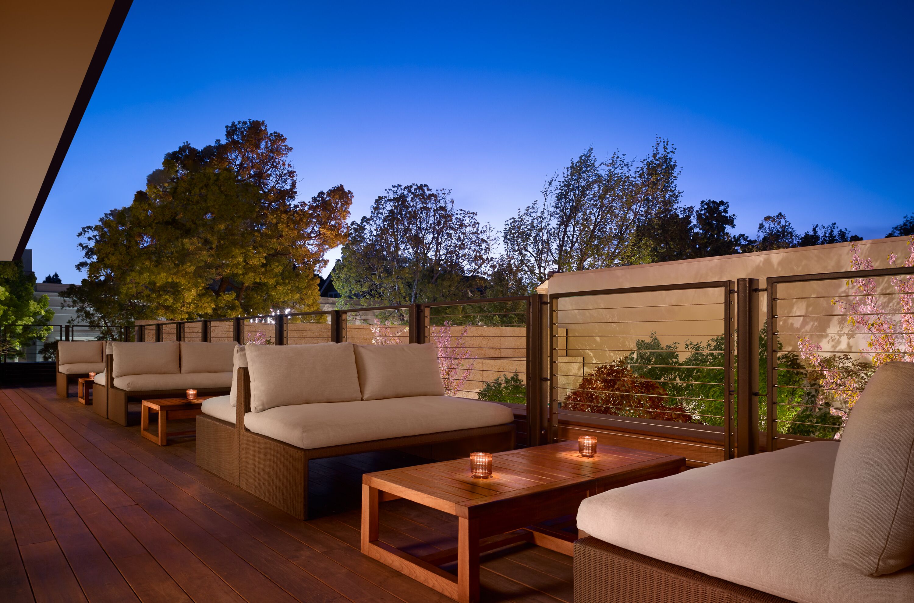 An outdoor patio with seating and tables, surrounded by a wooden deck and glass walls offering views of trees and a garden.