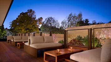 An outdoor patio with seating and tables, surrounded by a wooden deck and glass walls offering views of trees and a garden.