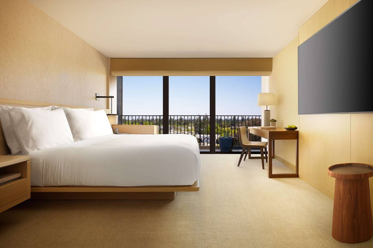 A modern hotel room with a large bed, a desk, a chair, a TV, and a balcony with a view.