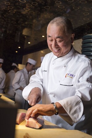 A chef in a white uniform with a logo, preparing food in a kitchen with other chefs in the background. - Nobu Hotel Palo Alto (Palo Alto)