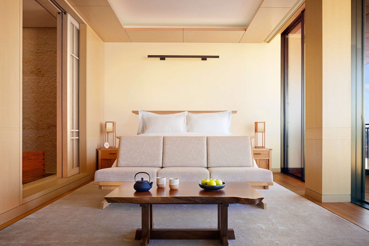Una habitación de hotel moderna con una cama grande, una mesa de centro de madera y vistas a la ciudad a través de las ventanas.