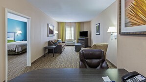 Quarto de hotel com cama, sofá, escrivaninha, cadeira, televisão e janela com cortinas.
