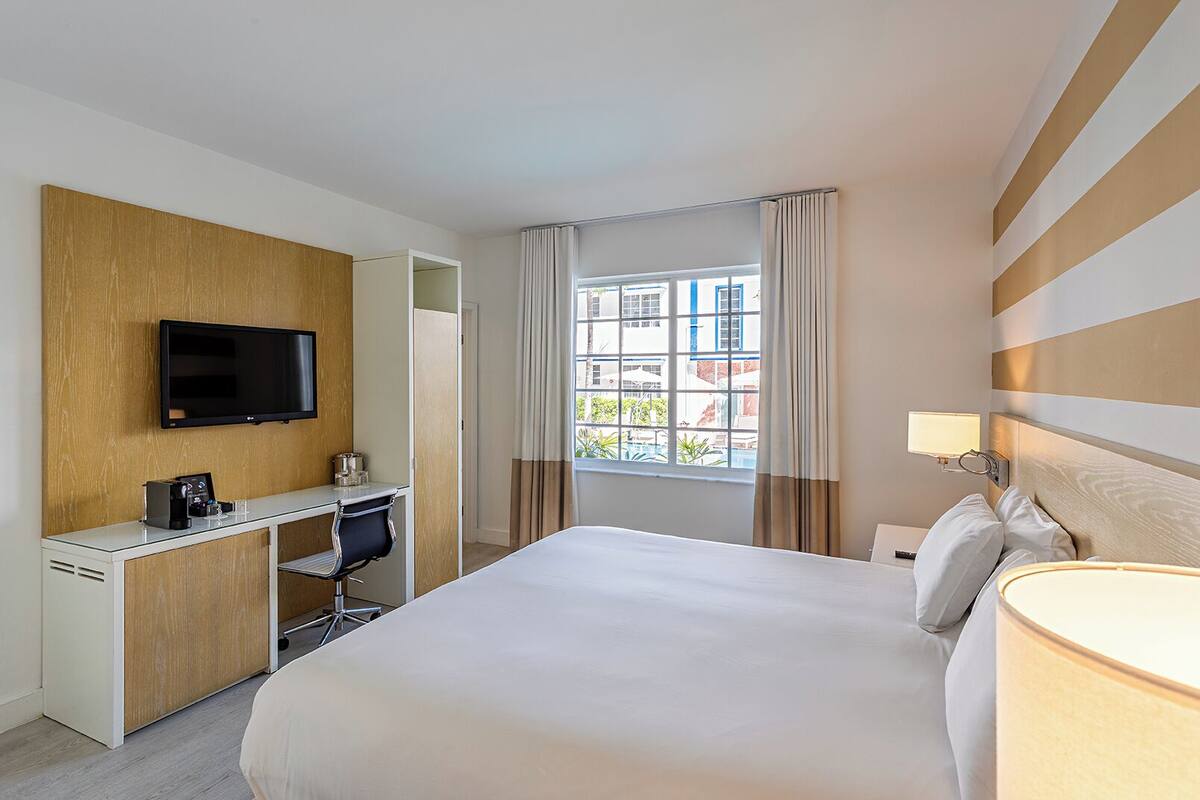 Ein Hotelzimmer mit einem Bett, einem Schreibtisch mit Stuhl, einem Fernseher und einem Fenster mit Vorhängen.