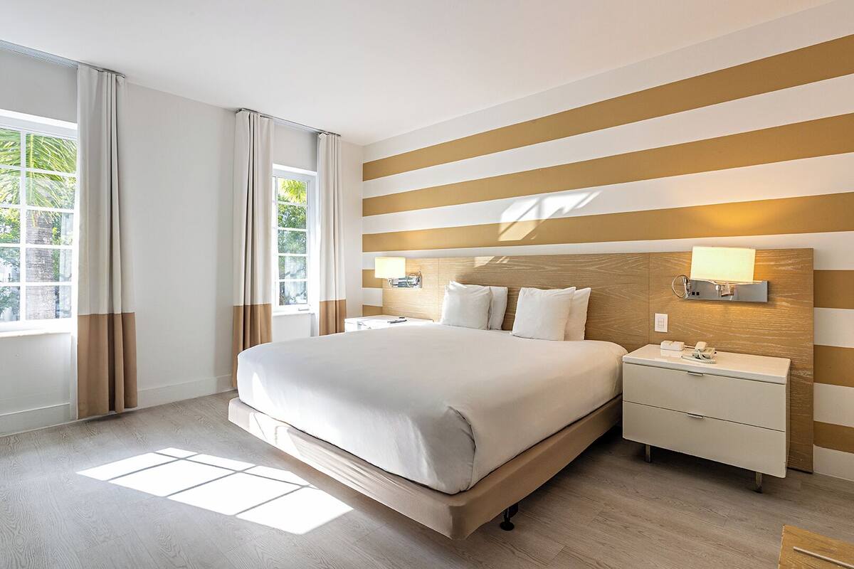 Ein Hotelzimmer mit einem großen Bett, Nachttischen, einer Wandlampe und einem Fenster mit Vorhängen.