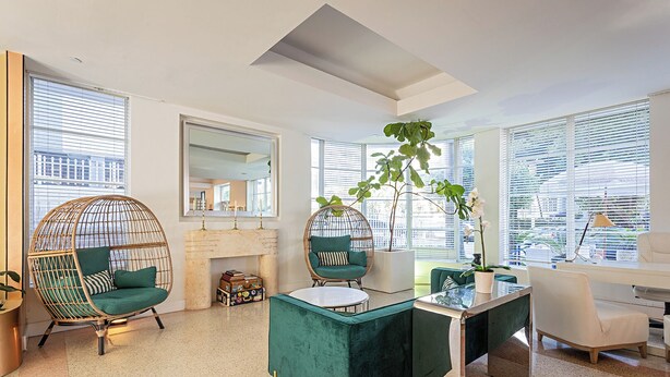 Un salon moderne avec une cheminée, deux canapés verts, un fauteuil rond en osier, un bureau et de grandes fenêtres.