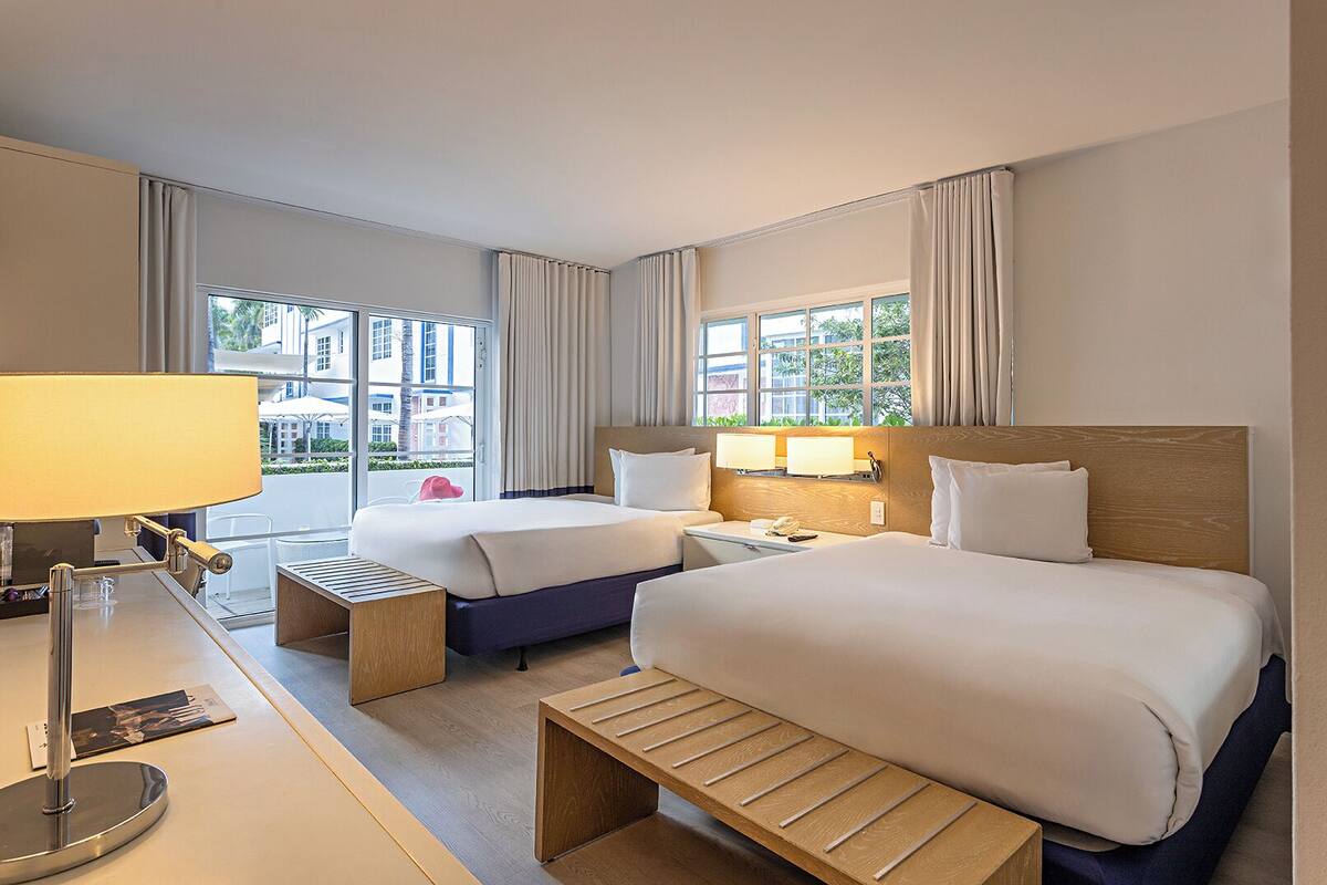 Ein Hotelzimmer mit zwei Betten, einem Schreibtisch mit Lampe, einem Nachttisch und einem Fenster mit Vorhängen.