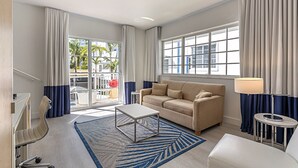 Een moderne hotelkamer met een beige bank, een salontafel, een televisie en uitzicht op palmbomen door grote ramen.
