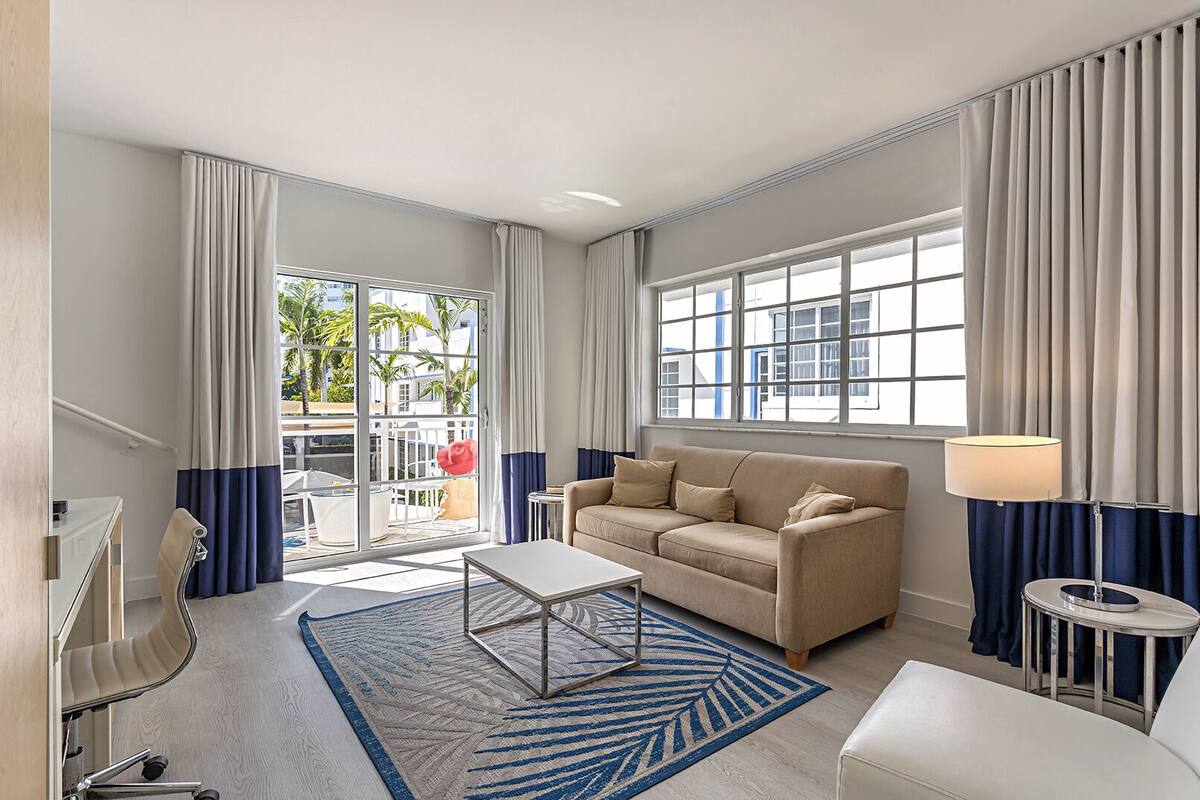 Ein modernes Hotelzimmer mit einem beige farbenen Sofa, einem Couchtisch, einem Fernseher und Blick auf Palmen durch große Fenster.