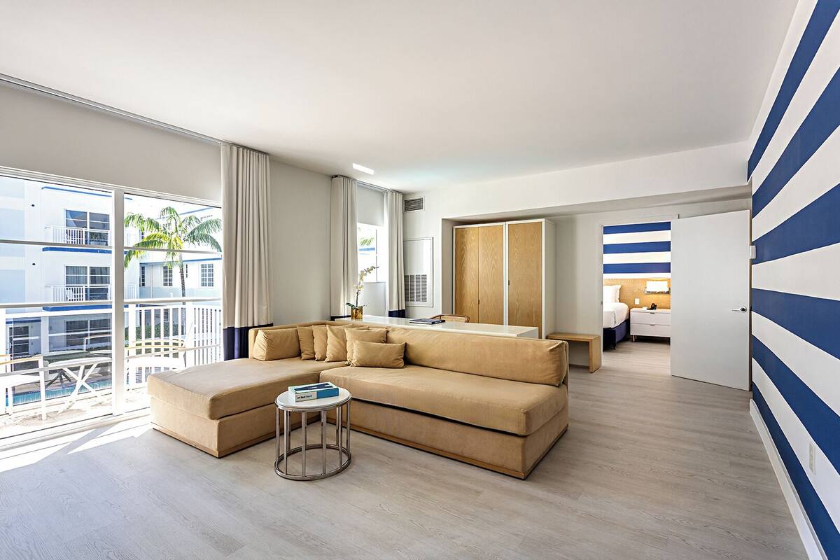 Ein modernes Hotelzimmer mit einer großen Couch, einem kleinen runden Tisch und Blick auf Pool und Gebäude.